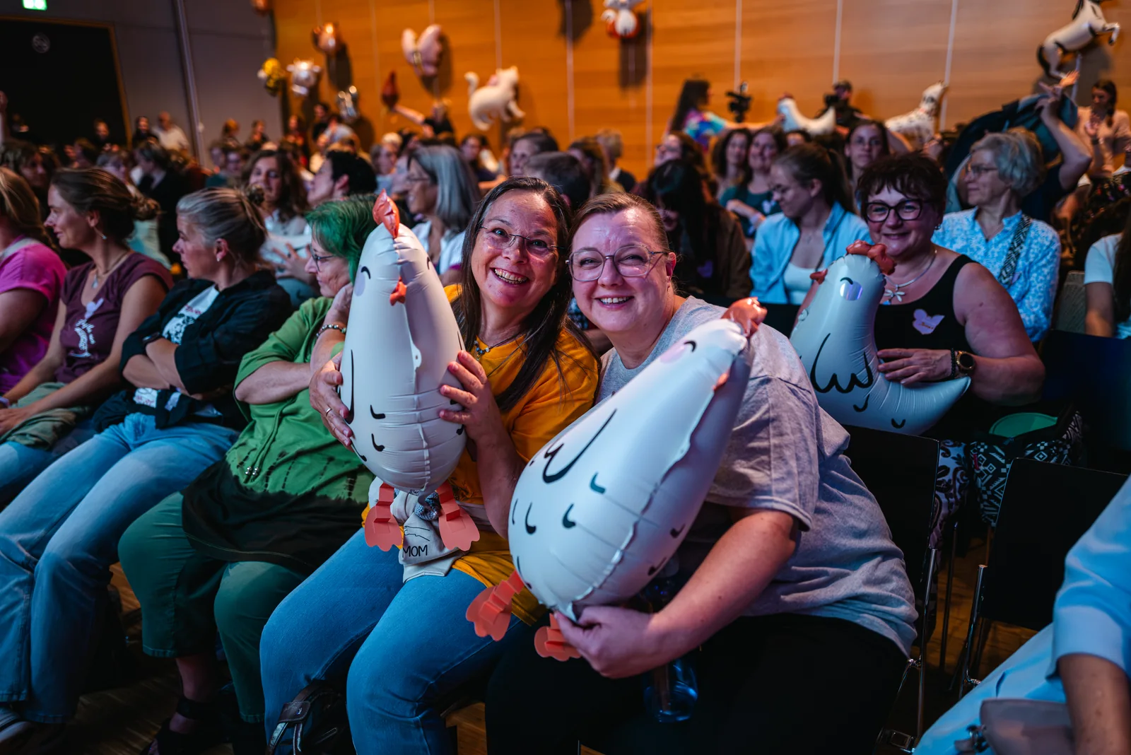 Zwei Freundinnen umarmen sich lachend mit Hühnerballons im Event-Publikum